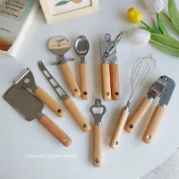 Set alat dapur gagang kayu minimalis berisi pengupas, parutan, pembuka botol, pengocok telur, dan alat serbaguna di atas meja putih dengan dekor bunga tulip.