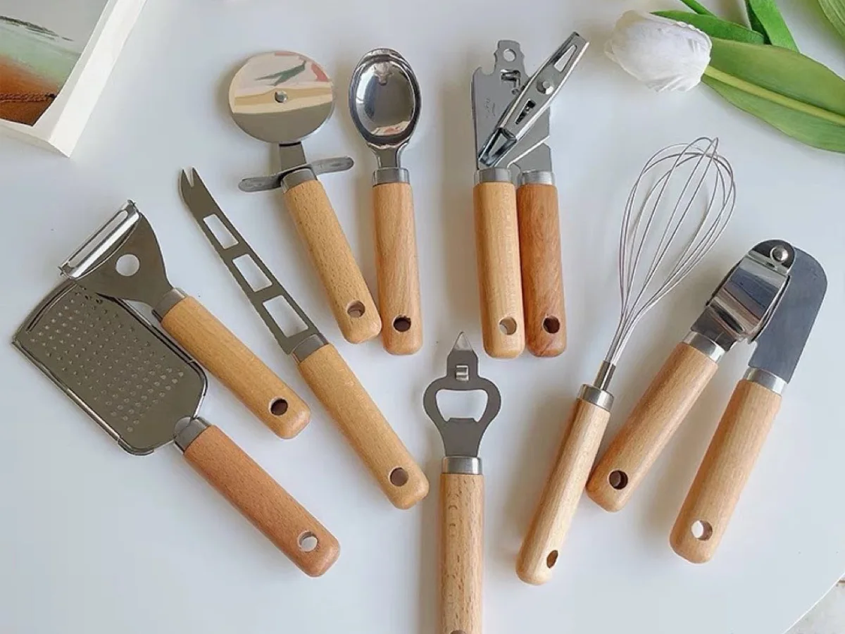 Set alat dapur gagang kayu minimalis berisi pengupas, parutan, pembuka botol, pengocok telur, dan alat serbaguna di atas meja putih dengan dekor bunga tulip.