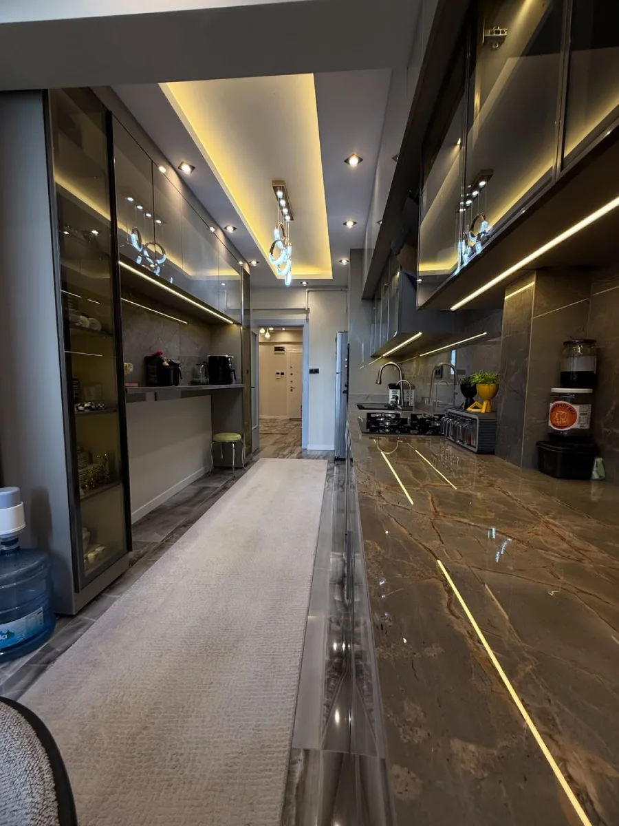 Lorong dapur apartemen mewah dengan meja marmer coklat berurat emas (gold veins) dan lemari penyimpanan kaca yang elegan.