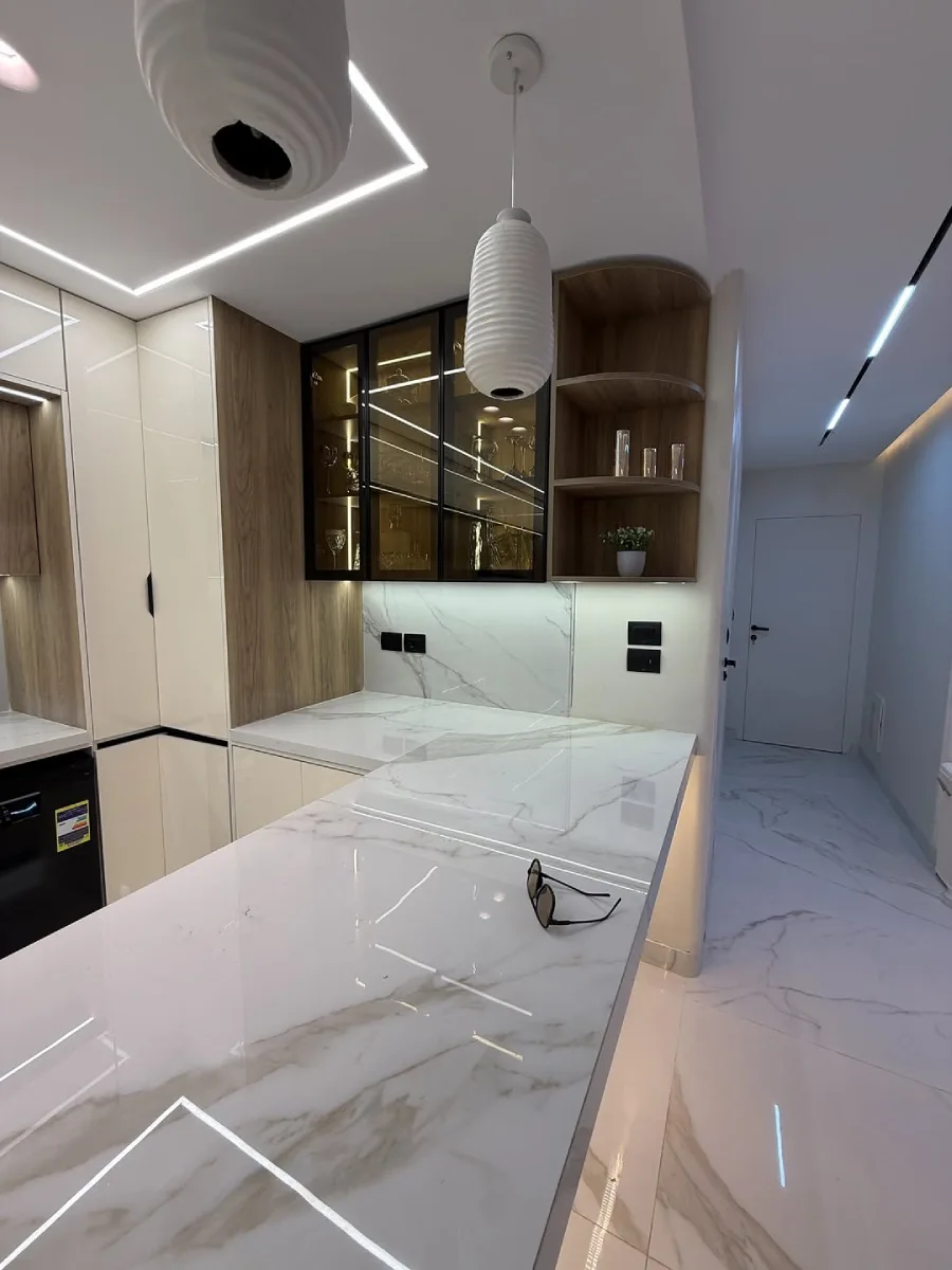 Meja island dapur marmer putih elegan dengan kabinet sudut melengkung dan sentuhan motif kayu natural.