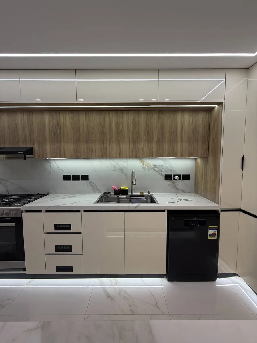 Area wastafel dapur bersih dengan kabinet bawah warna beige muda, mesin pencuci piring hitam, dan backsplash marmer statuario.