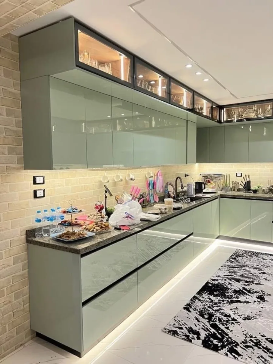 Kitchen set modern letter L finishing glossy warna hijau sage muda dengan aksen dinding bata ekspos krem dan pencahayaan LED strip bawah kabinet.