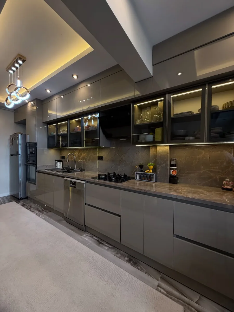 Kitchen set mewah bernuansa abu-abu taupe high-gloss dengan lemari pajangan kaca (glass display cabinet) dan lampu gantung cincin modern.