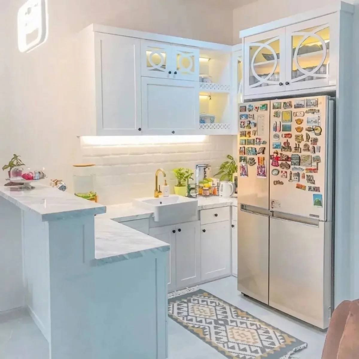 Dapur minimalis elegan serba putih dengan sentuhan meja marmer, keran emas (gold faucet), dan kabinet kaca dengan pencahayaan dalam (ambient lighting).