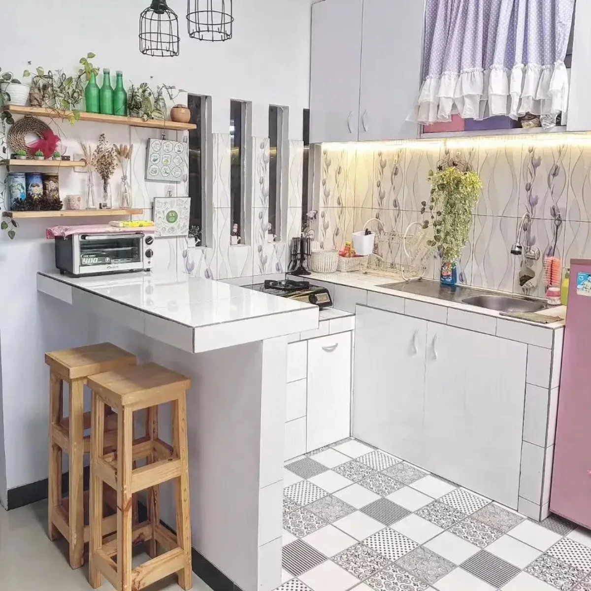 Dapur minimalis mungil dengan meja sarapan (breakfast bar) keramik putih, kursi kayu tinggi, dan sentuhan dekorasi nuansa ungu pastel serta rak dinding ambalan.