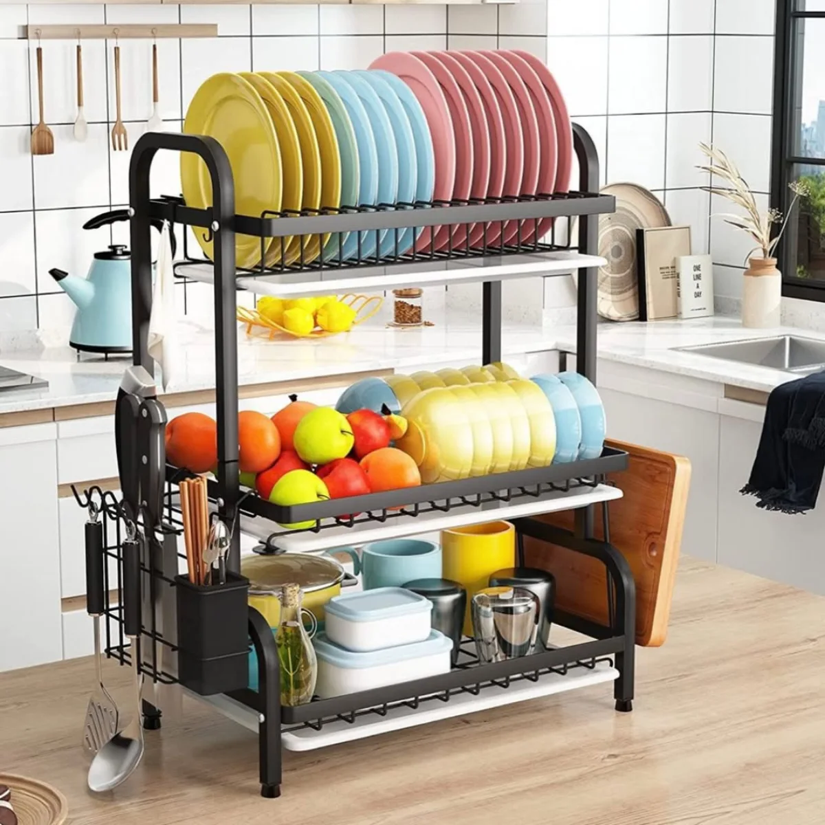 Rak piring modern berwarna hitam di atas meja dapur berisi susunan piring warna-warni, mangkuk, buah-buahan segar, dan tempat alat makan