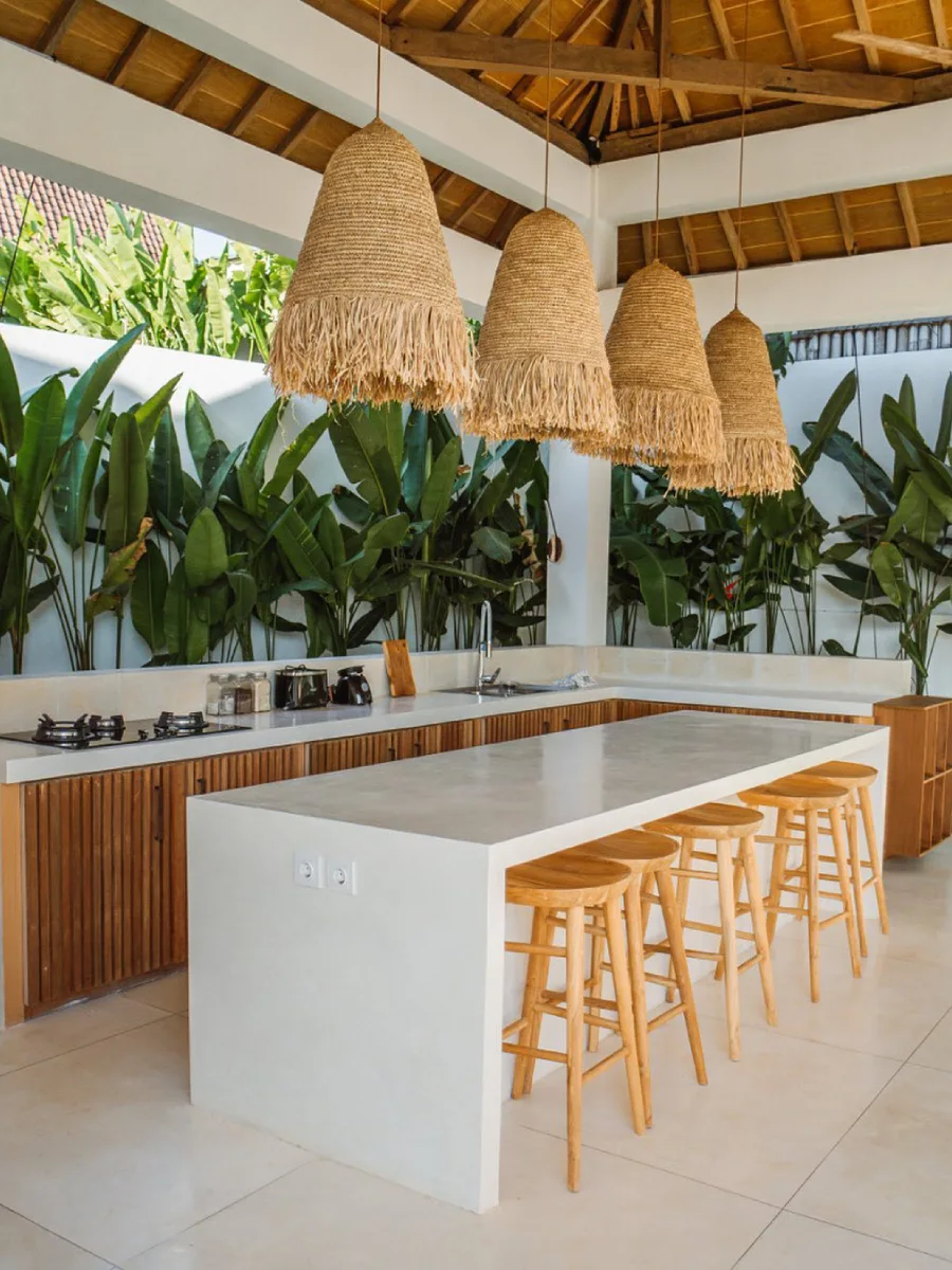 Dapur semi-outdoor gaya vila tropis dengan meja island beton putih minimalis, lampu gantung rotan boho berukuran besar, dan latar belakang tanaman hijau rimbun.