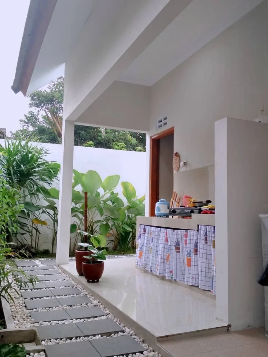 Desain dapur kotor di teras belakang (backyard kitchen) yang menyatu dengan taman, dilengkapi meja cor beton, tirai kolong motif kotak-kotak, dan jalan setapak batu koral.