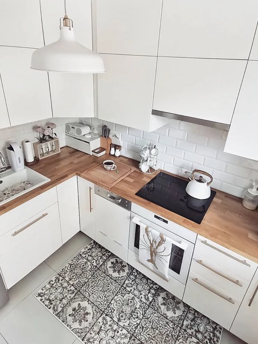 Layout dapur bentuk L bergaya modern putih bersih dengan countertop kayu solid, wastafel keramik, backsplash bata putih, dan lantai dengan karpet motif vintage.