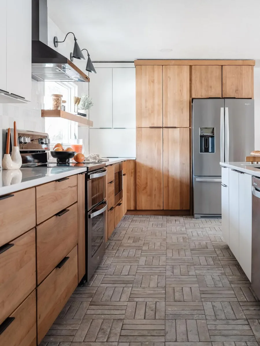 Dapur luas dengan dominasi kabinet kayu natural hangat (warm wood), lantai keramik motif anyaman bata, kulkas side-by-side, dan kitchen island putih.