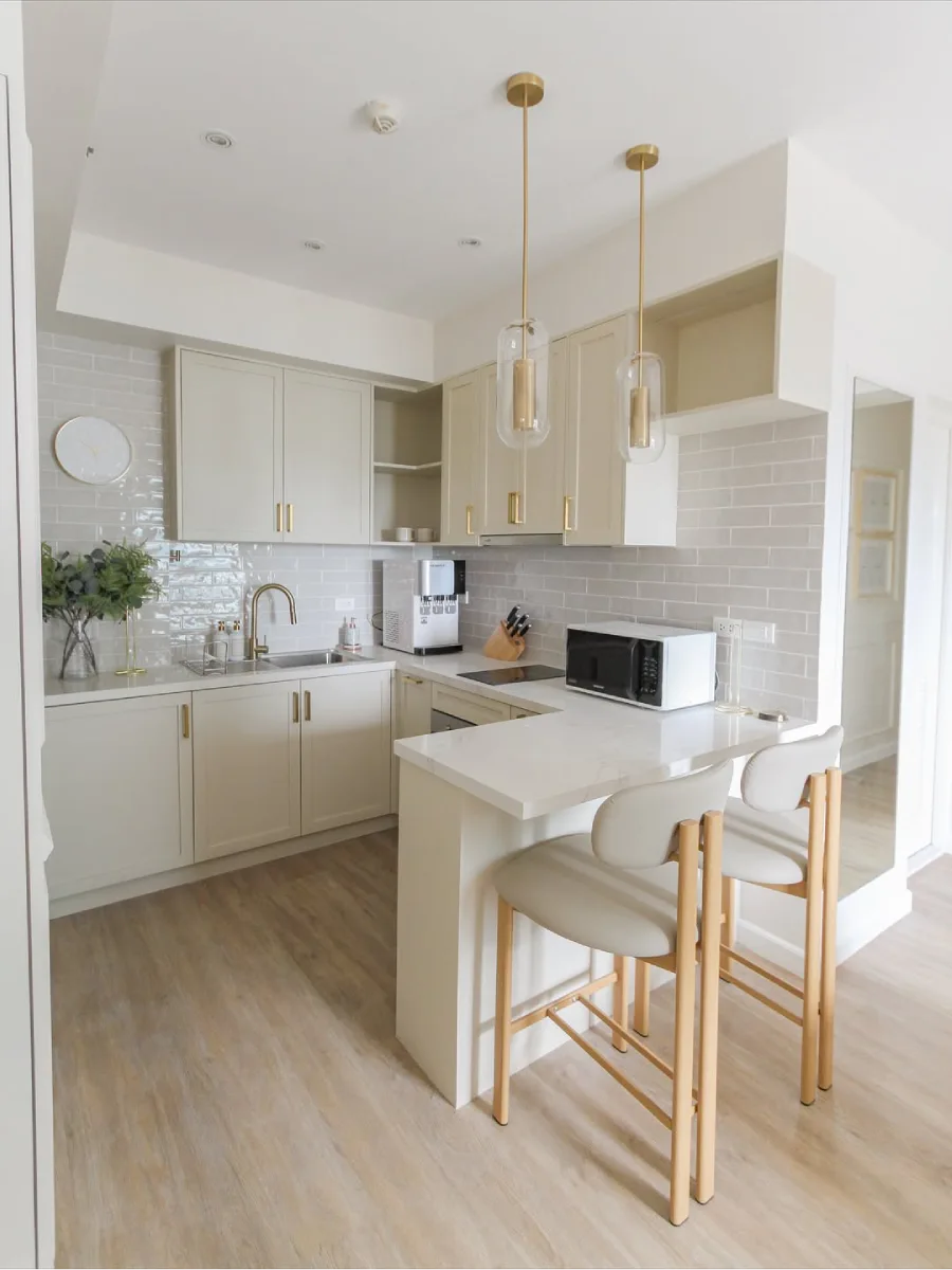 Dapur apartemen modern bernuansa luxury lembut, meja bar sarapan (minibar) menyatu dengan kabinet, lampu gantung kaca aksen emas, dan kursi bar tinggi yang stylish.