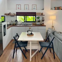 Meja makan lipat kayu berwarna terang berbentuk persegi dengan dua kursi lipat hitam di area dapur bernuansa putih dan kayu