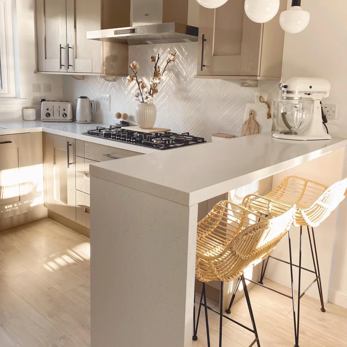 Meja bar semenanjung dapur berwarna putih bersih dengan desain waterfall edge yang dilengkapi dua kursi bar rotan di dapur yang cerah