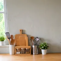 Dinding dapur kusam berwarna abu-abu terang dengan perlengkapan dapur sederhana di atas meja kayu dan pencahayaan alami dari jendela.