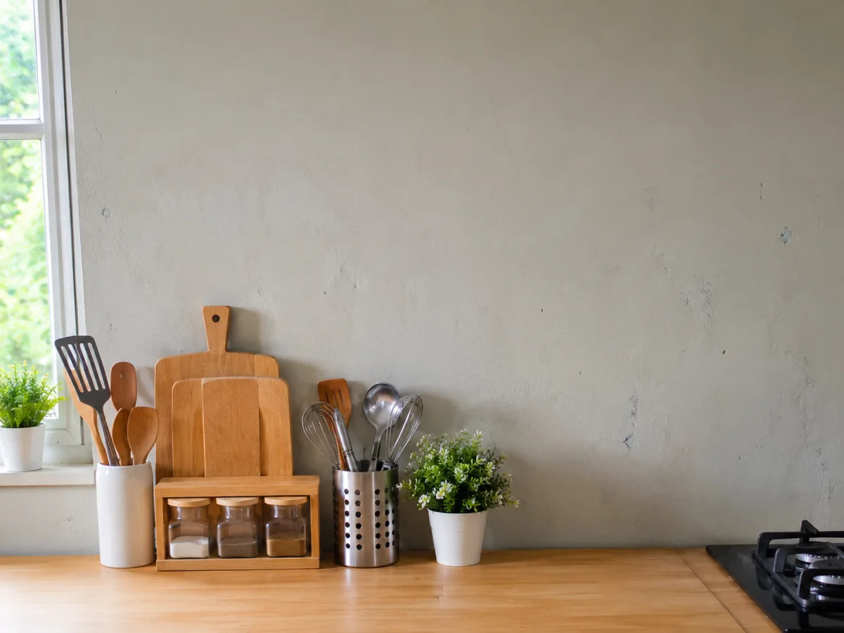Dinding dapur kusam berwarna abu-abu terang dengan perlengkapan dapur sederhana di atas meja kayu dan pencahayaan alami dari jendela.