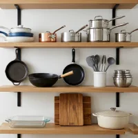 Rak dapur kayu terapung empat tingkat yang ditata rapi menampilkan berbagai peralatan masak seperti wajan keramik, panci stainless steel dan tembaga, wok, alat silikon, mangkuk, loyang kaca, talenan kayu, dan Dutch oven, dengan latar belakang dapur minimalis yang cerah.