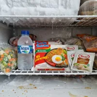 Tampak dalam kompartemen freezer dengan es yang mencair dan air menetes membasahi kemasan makanan seperti Indomie dan botol Aqua, menunjukkan kondisi akibat kulkas tidak dingin.