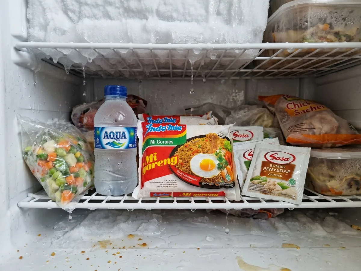 Tampak dalam kompartemen freezer dengan es yang mencair dan air menetes membasahi kemasan makanan seperti Indomie dan botol Aqua, menunjukkan kondisi akibat kulkas tidak dingin.