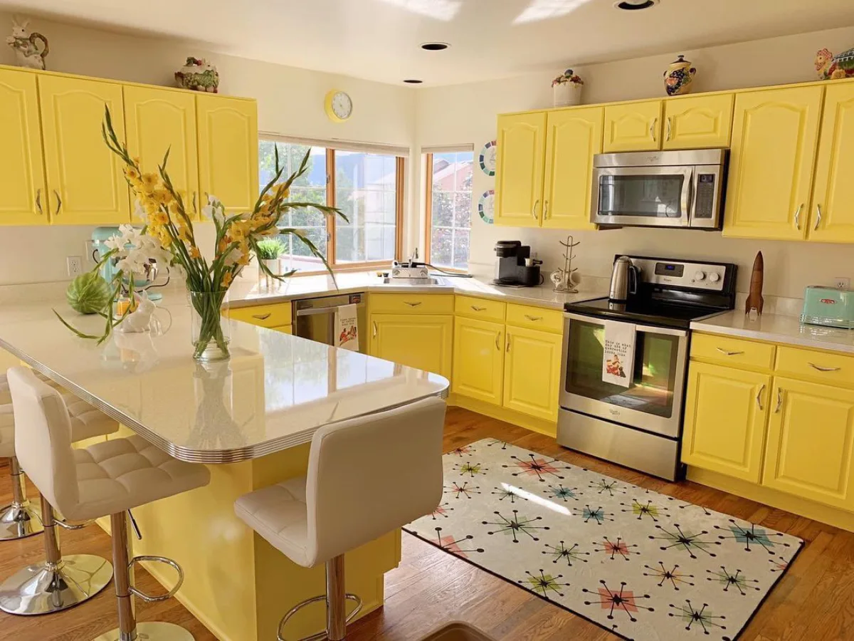 Dapur luas bertema retro dengan kabinet warna kuning kenari yang mencolok, lantai kayu, dan meja island besar berwarna putih.