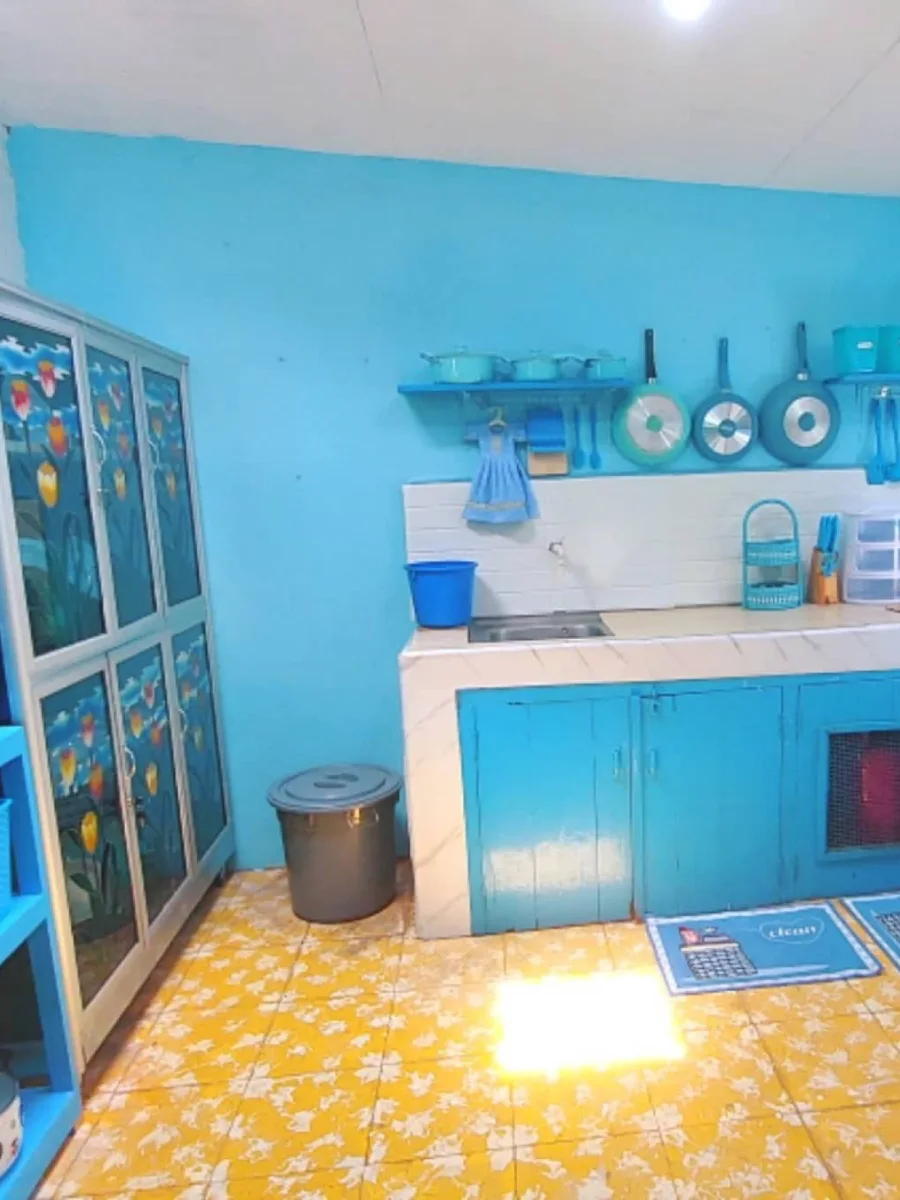Sisi lain dapur berwarna biru laut terang dengan lemari kaca besar berstiker bunga, ember penyimpanan, dan peralatan masak gantung berwarna senada.