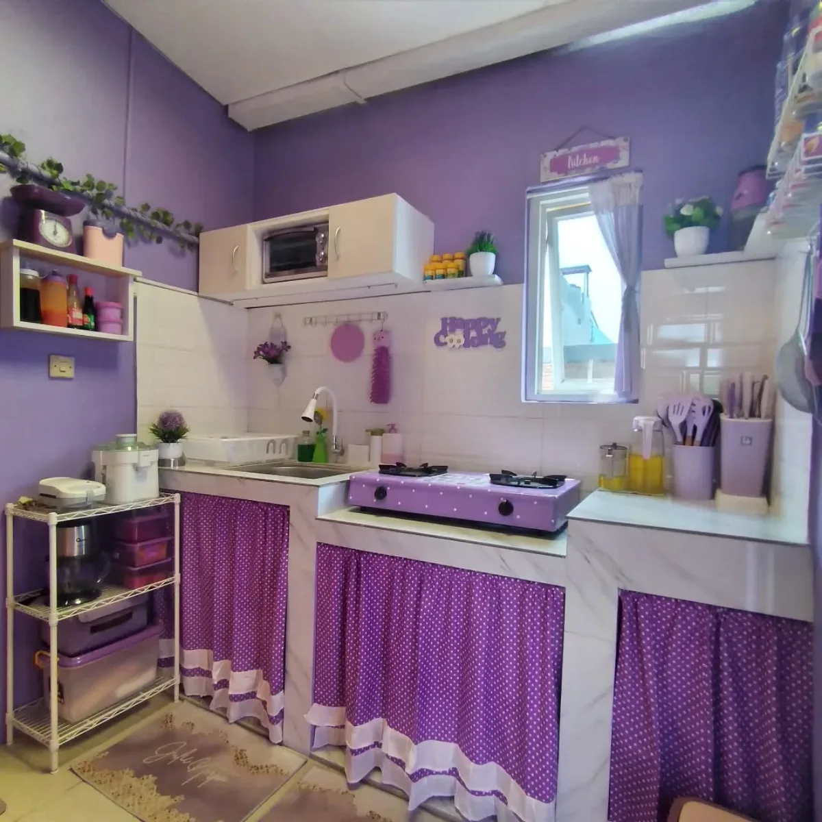 Layout dapur lurus (linear kitchen) warna ungu pastel dengan pencahayaan alami dari jendela kecil dan perabotan serba ungu.