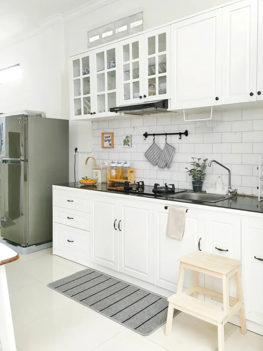Model kitchen set minimalis warna putih duco dengan kabinet atas pintu kaca, top table granit hitam, dan backsplash bata putih (subway tile).