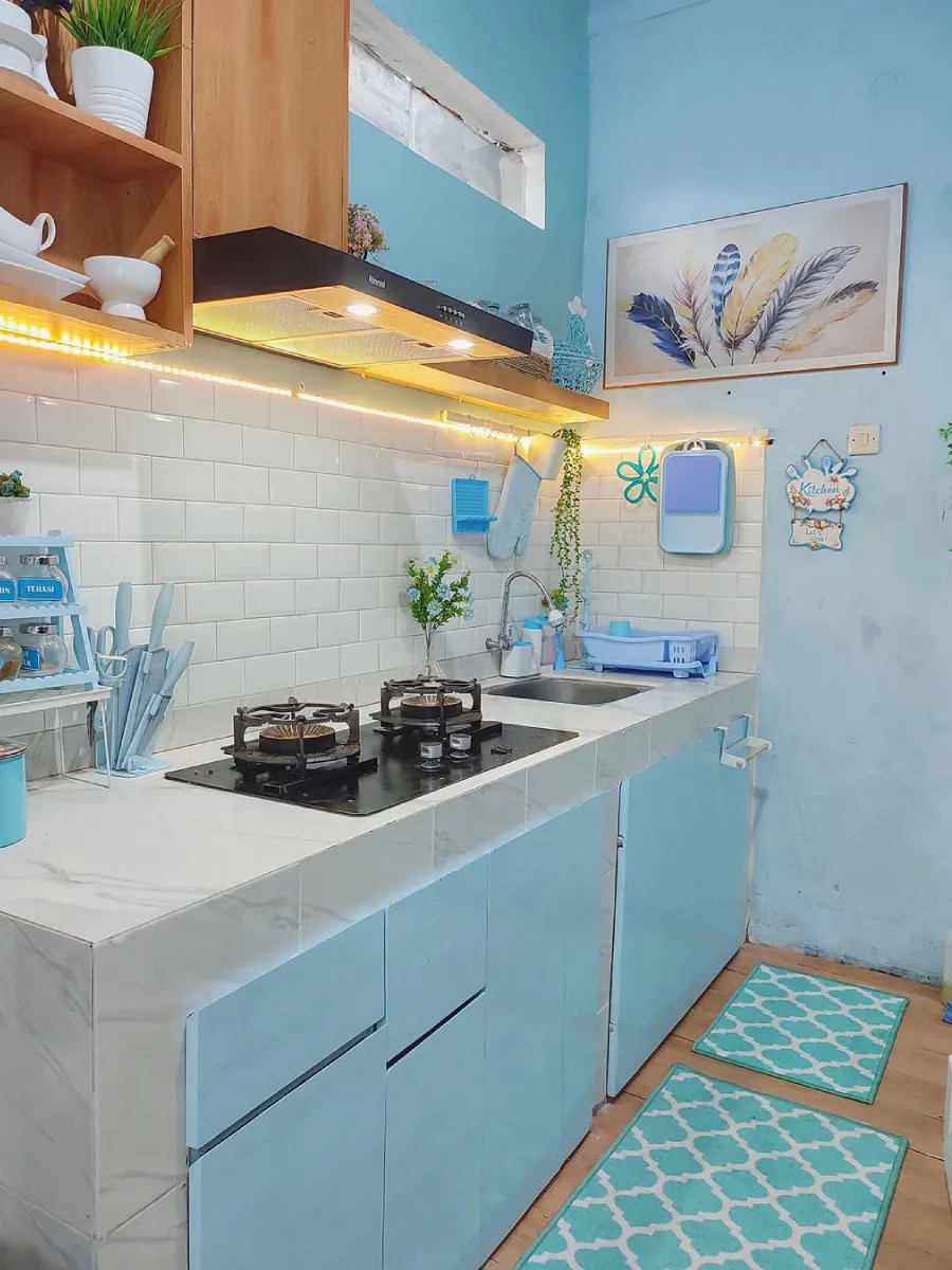 Area wastafel dapur dengan kabinet bawah berwarna biru langit lembut, backsplash keramik bata putih, dan rak kayu terbuka dengan pencahayaan LED hangat.
