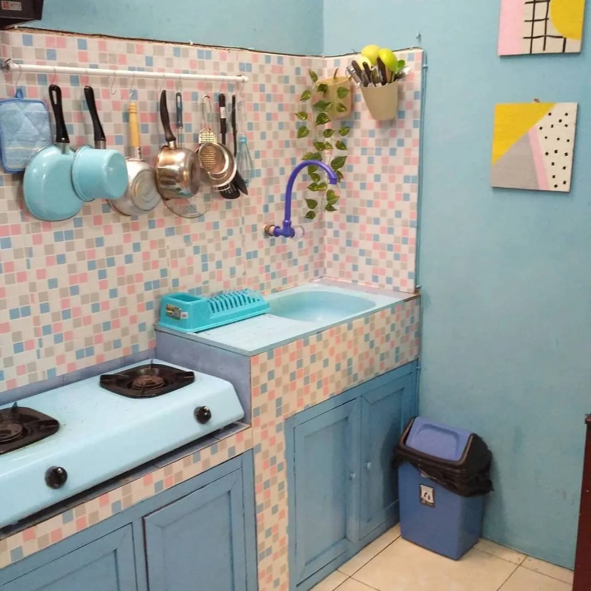 Sudut dapur mungil dengan kabinet bawah cat warna dusty blue, dinding keramik mosaik warna-warni pastel, dan peralatan masak gantung yang rapi.