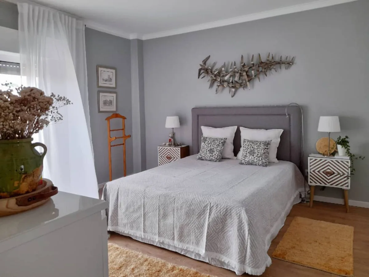 Interior kamar tidur modern dengan cat dinding abu-abu terang, hiasan dinding logam artistik, dan aksen karpet kuning.