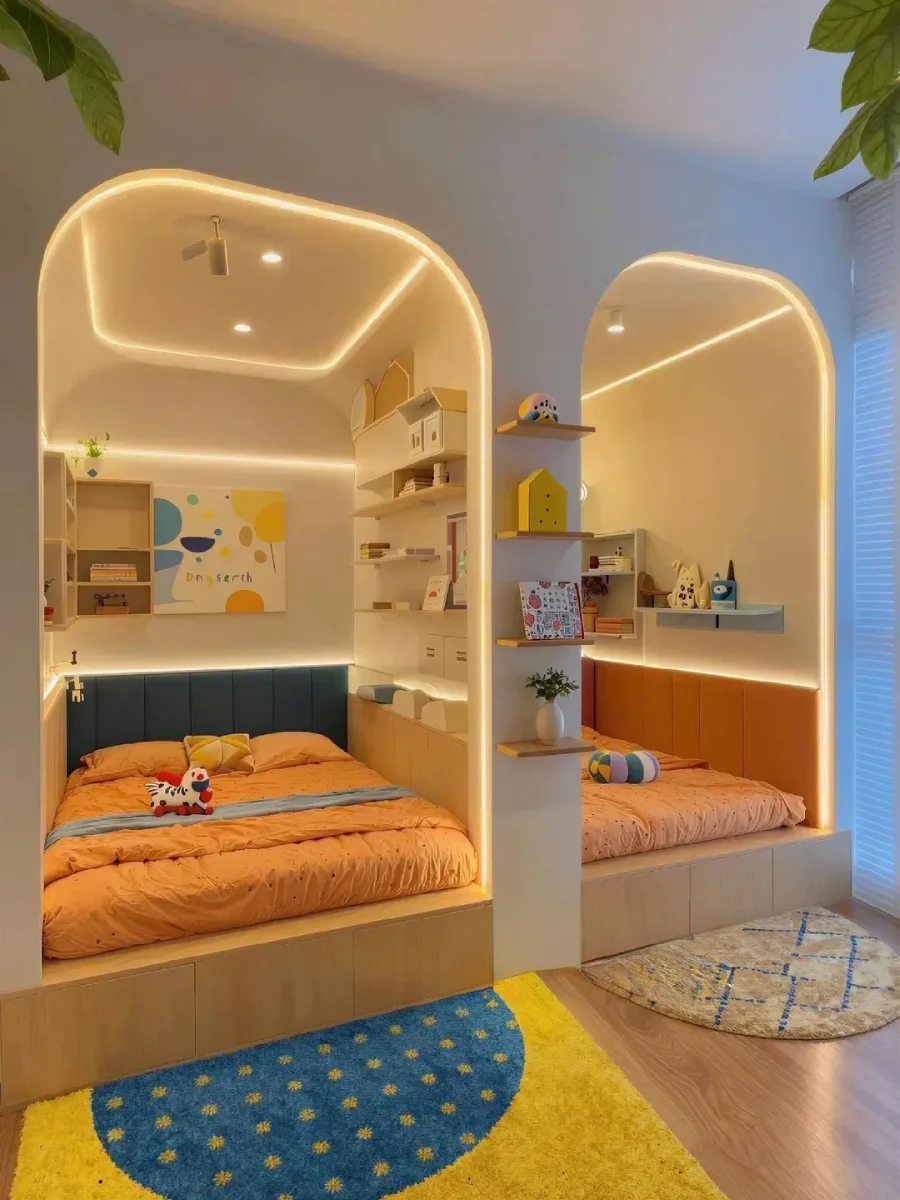 Pemandangan interior kamar tidur anak kembar dengan dua ceruk (nook) tempat tidur melengkung berdampingan. Masing-masing memiliki pencahayaan LED tersembunyi di sepanjang tepi, tempat tidur platform kayu dengan laci penyimpanan, dan rak gantung terbuka. Lantai kayu ditutupi karpet berwarna-warni.