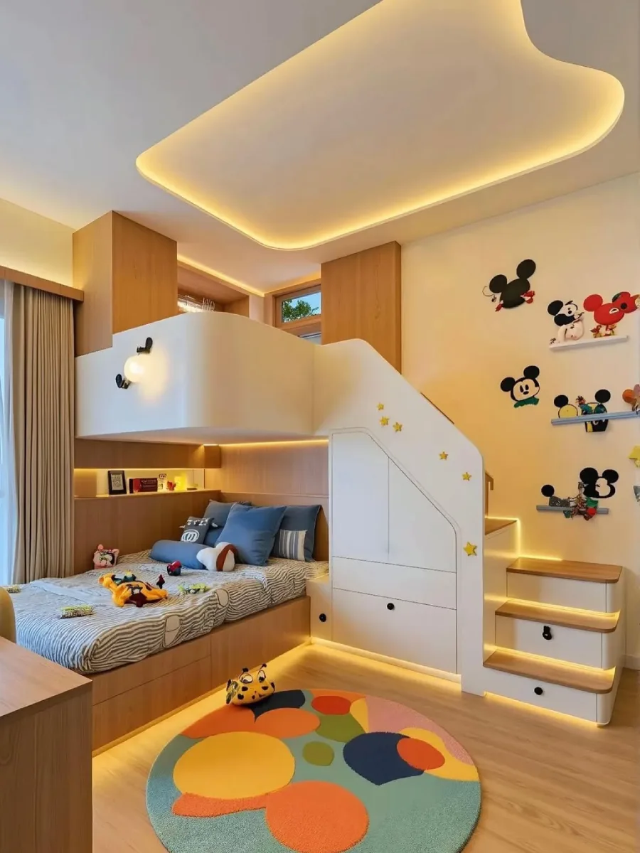 Kamar tidur anak modern yang cerah dengan tempat tidur susun terintegrasi, tangga putih dengan detail bintang, area belajar, mural dinding Mickey Mouse, dan karpet bundar berwarna-warni di bawah langit-langit beraksen cahaya.