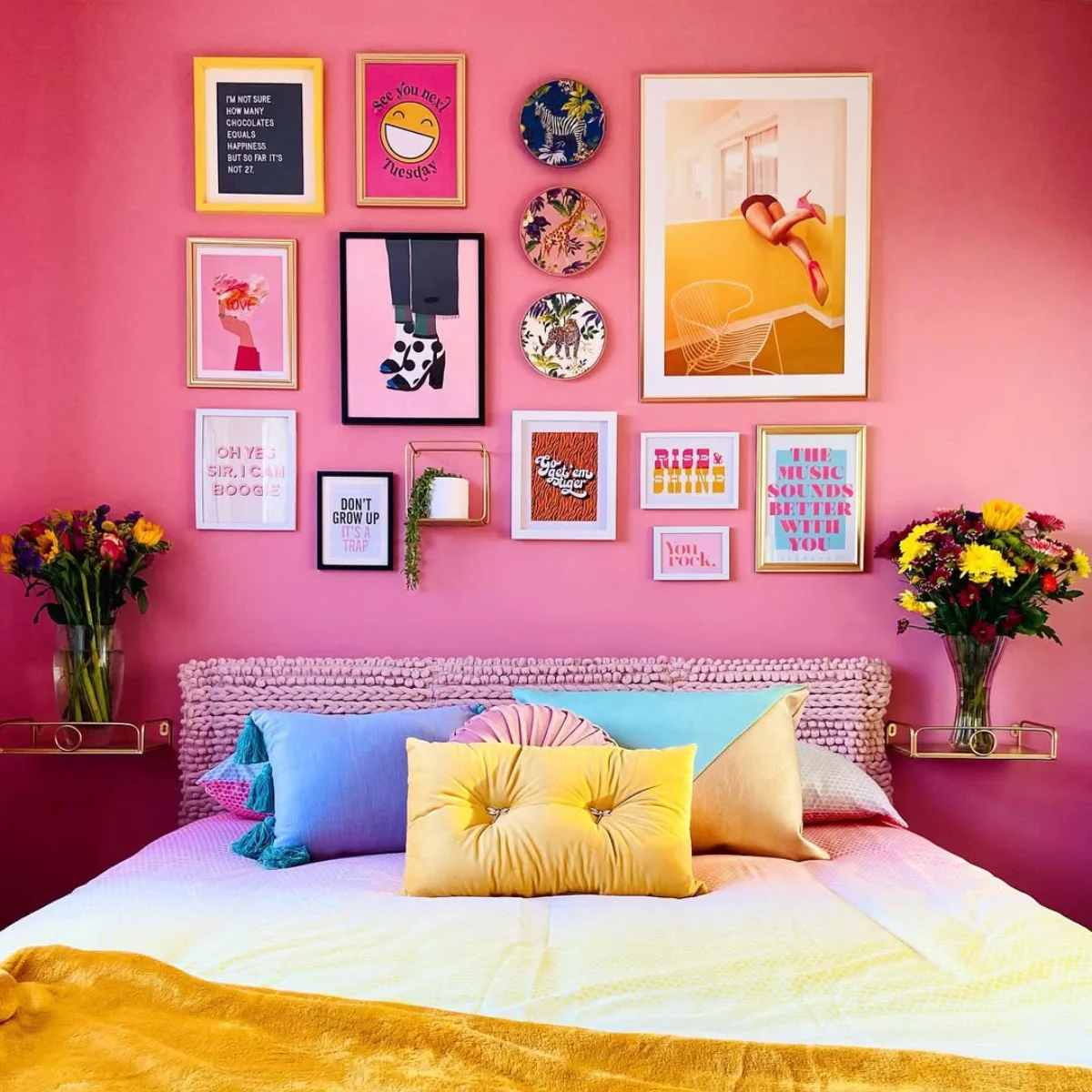 Susunan eklektik bingkai gambar, poster art print dengan cetakan sepatu dan tipografi quotes, serta piring hias melingkar di dinding kamar tidur berwarna merah muda di atas tempat tidur dengan bantal-bantal warna-warni.