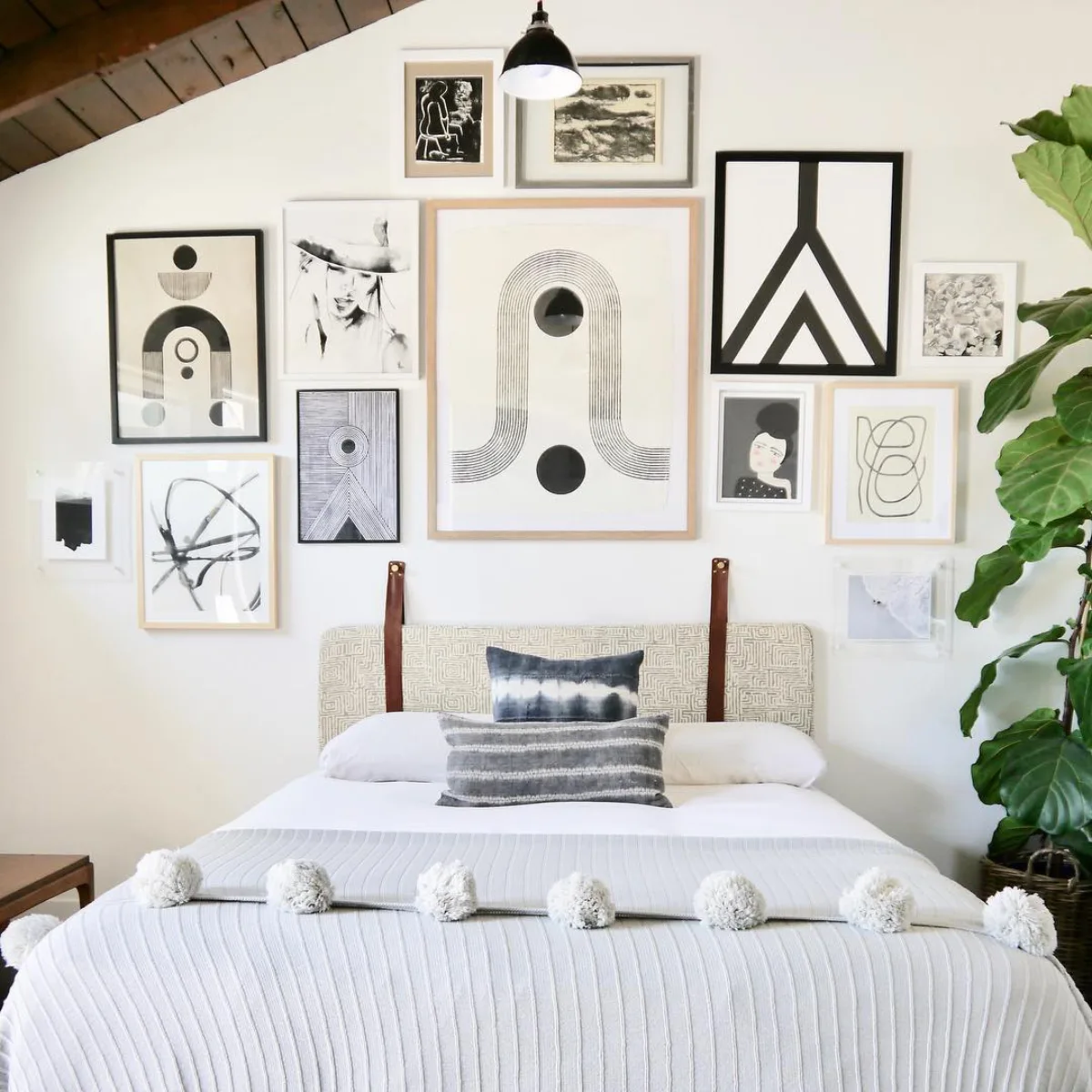 Galeri dinding bergaya monokrom minimalis yang berisi perpaduan seni abstrak hitam putih dan line art di atas tempat tidur putih berselimut pom-pom