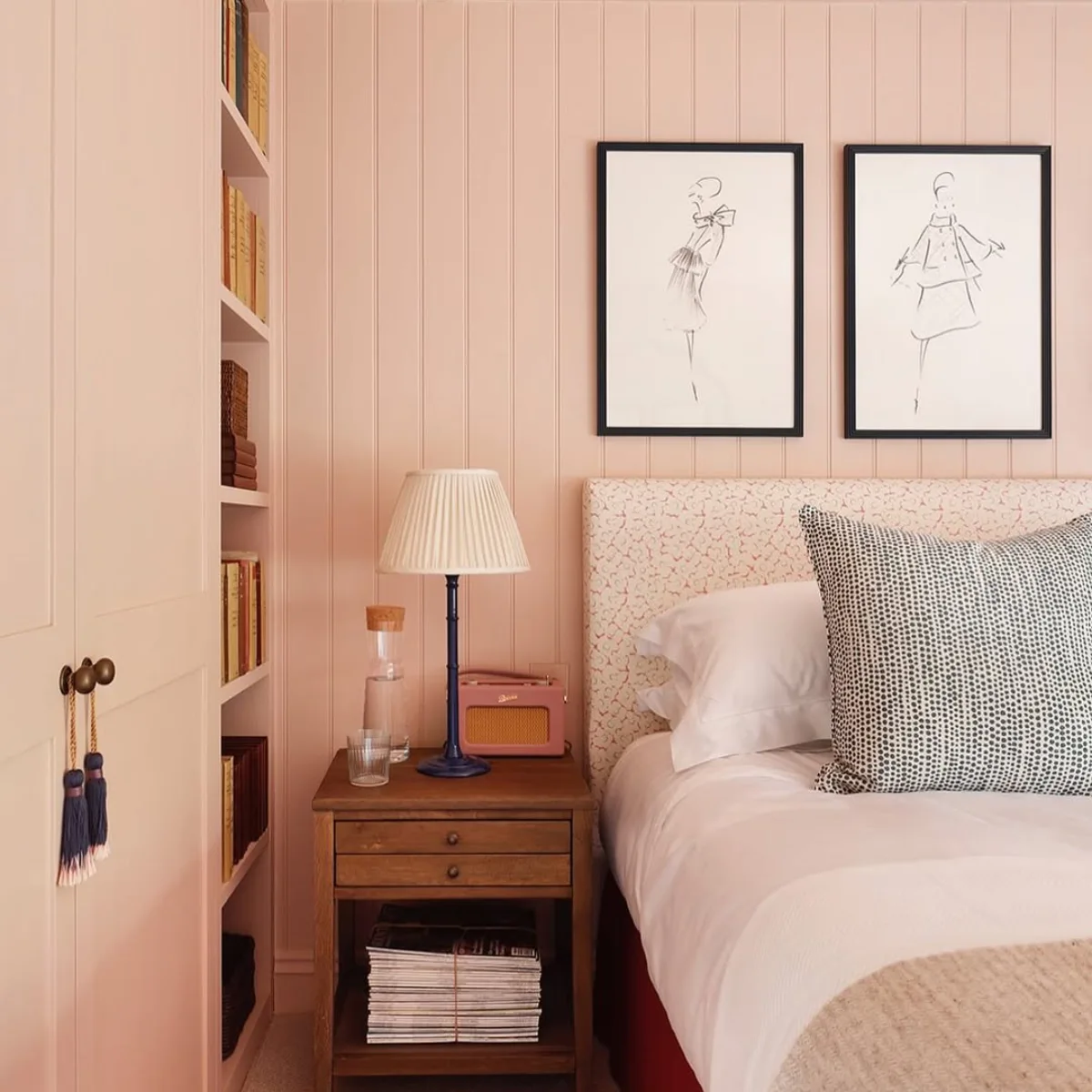 Sudut kamar tidur estetik bernuansa pink pastel dengan dinding panel kayu, rak buku built-in, nakas kayu klasik dengan radio vintage, dan dua sketsa fashion hitam putih di dinding.