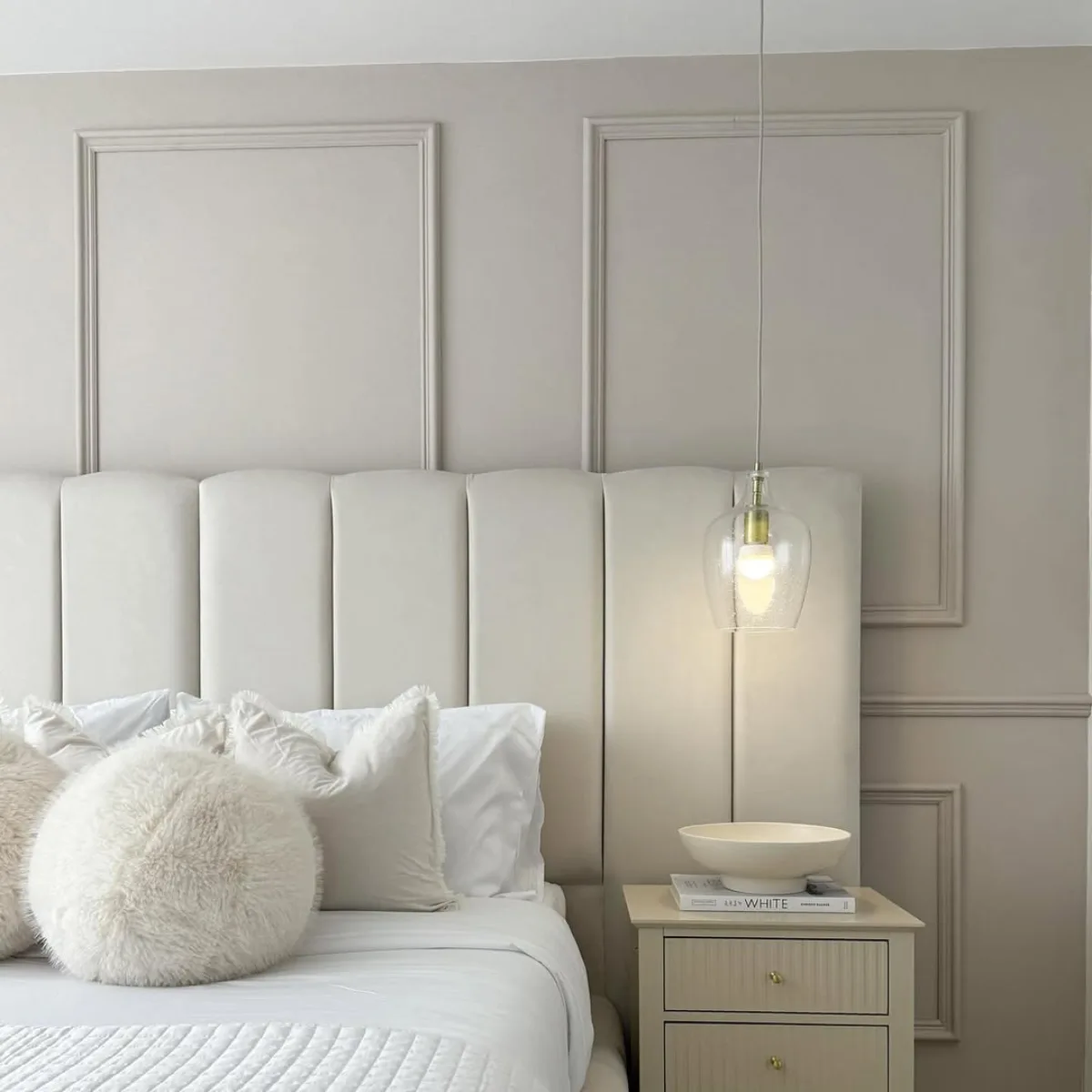 Detail interior kamar tidur elegan dengan headboard panel empuk warna beige, lampu gantung kaca modern, bantal dekoratif bulu bulat, dan nakas samping tempat tidur tiga laci.