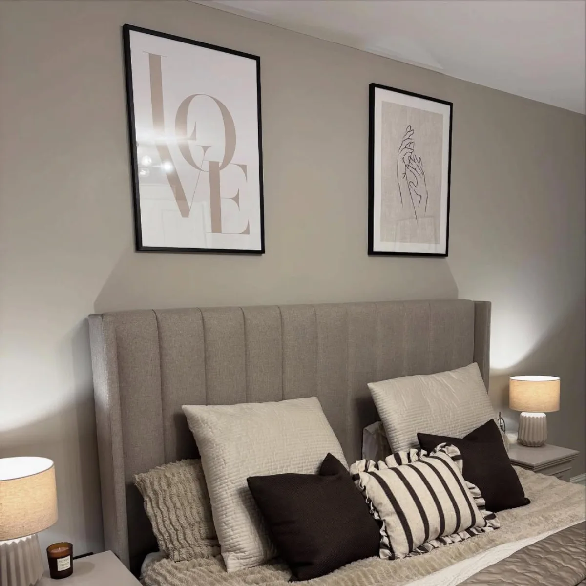 Tempat tidur mewah dengan headboard sandaran tinggi berlapis kain abu-abu, bantal aksen warna coklat dan garis-garis, serta sepasang lampu meja di atas nakas klasik.