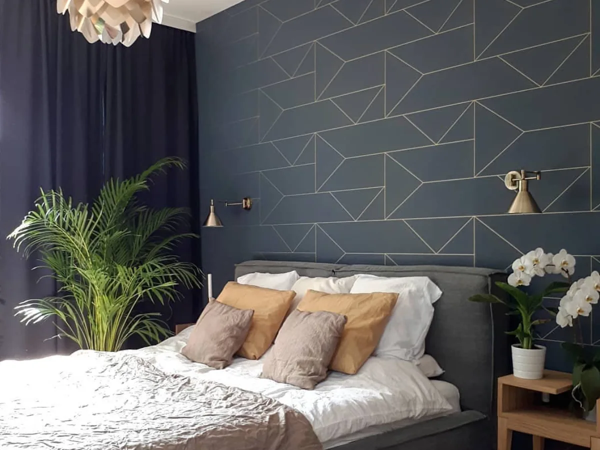 Kamar tidur elegan dengan dinding cat biru dongker keabu-abuan bermotif garis geometris, dipadukan tempat tidur abu-abu, sprei netral, dan pencahayaan lembut.