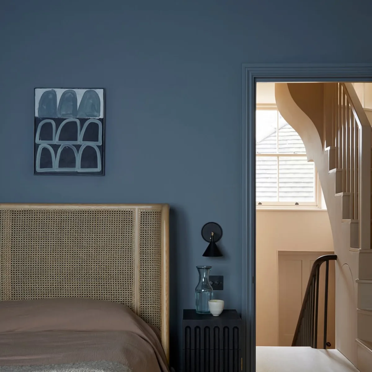 Kombinasi warna cat dinding slate blue atau biru keabu-abuan yang sejuk dengan tempat tidur sandaran rotan alami dan meja nakas hitam modern.