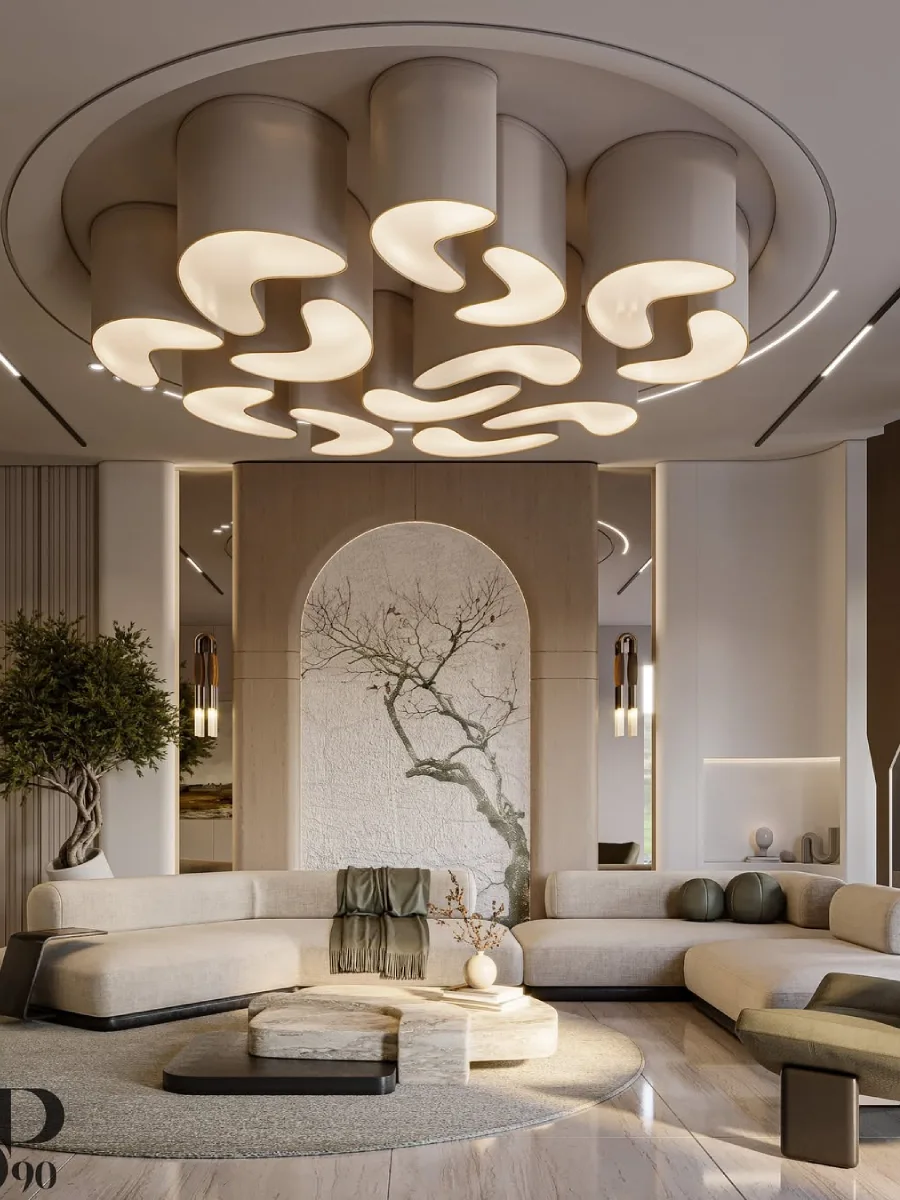 Ruang keluarga dengan konsep organik dan kurva yang dominan. Menampilkan sofa melengkung berwarna off-white dan meja kopi batu alam. Fokus utamanya adalah lampu plafon unik berbentuk pola lengkungan yang saling terkait dan dinding aksen dengan lukisan pohon bergaya oriental di dalam bingkai arkade.