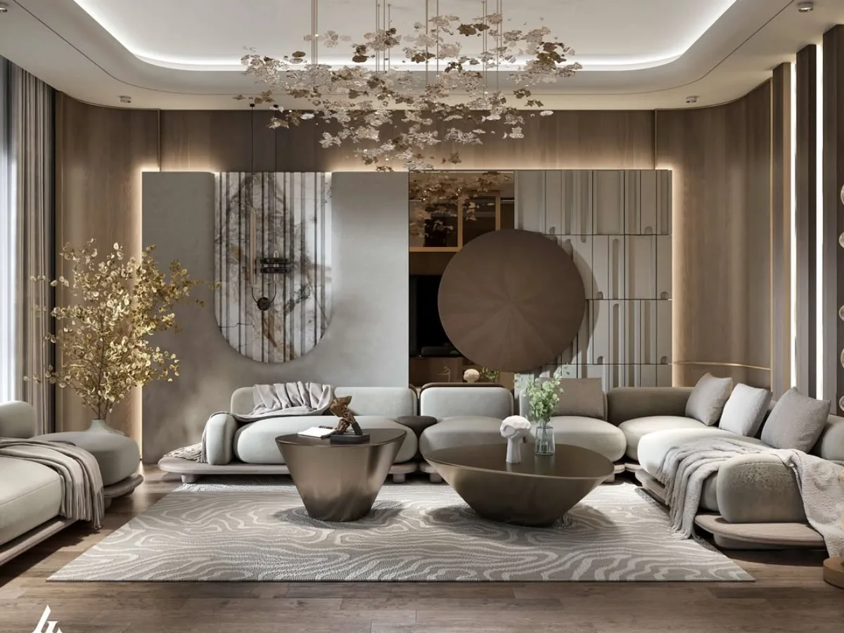 Interior ruang keluarga mewah dengan konsep modern kontemporer yang didominasi elemen kayu dan pencahayaan hangat. Fokus utama pada sofa modular berwarna abu-abu muda yang melengkung, dua meja kopi logam berbentuk asimetris dengan finishing perunggu, dan karpet bermotif abstrak bergelombang. Latar belakang menampilkan panel dinding kayu dengan aksen marmer vertikal serta lampu gantung artistik berbentuk kepingan emas yang menjuntai dari langit-langit tray ceiling.