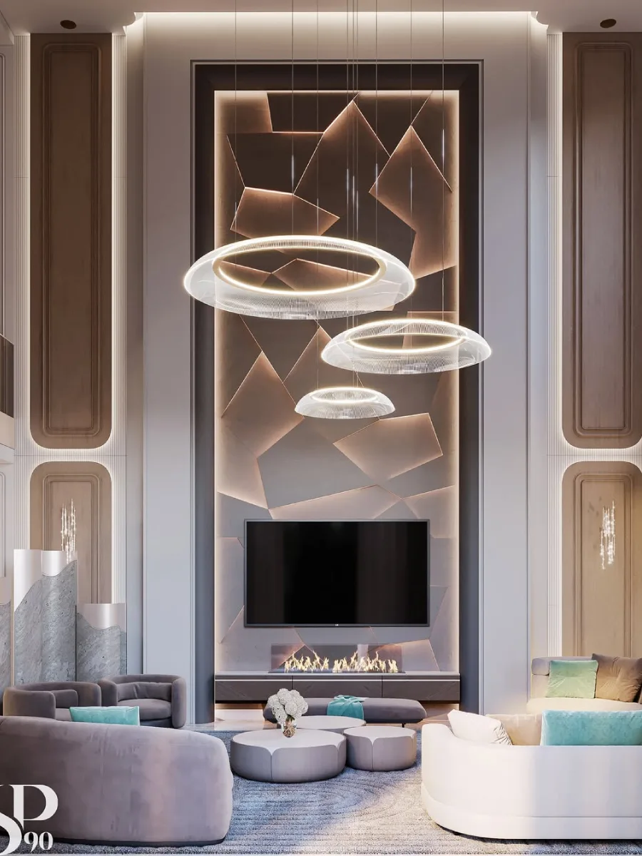 Ruang keluarga modern yang menampilkan dinding TV dengan panel geometris asimetris yang diterangi lampu LED tersembunyi. Ruangan ini dilengkapi dengan sofa melingkar berwarna abu-abu muda, aksen bantal tosca, dan lampu gantung minimalis berbentuk cincin cahaya yang tumpang tindih.