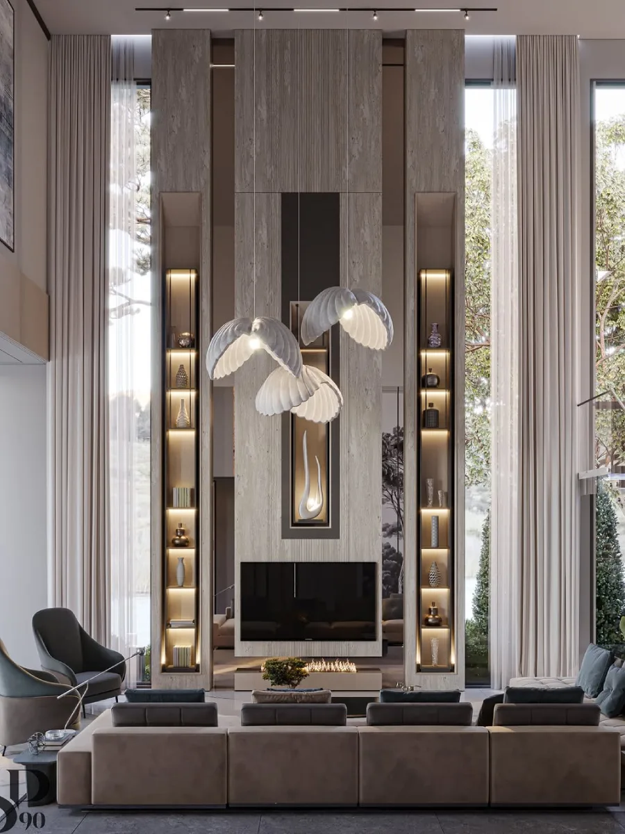 Desain ruang keluarga terbuka yang sangat tinggi dengan pemandangan taman melalui jendela kaca lebar. Fokus utama adalah dinding perapian dari batu alam abu-abu dengan rak buku ramping di kedua sisinya. Lampu gantung unik berbentuk seperti sayap burung atau kerang putih menghiasi bagian tengah ruangan.