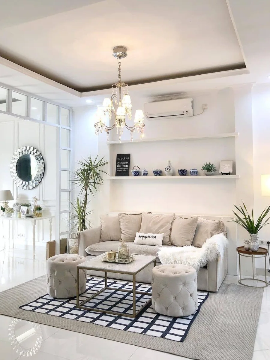Ruang keluarga serba putih yang elegan dengan lampu gantung kristal, ottoman beludru, dan rak pajangan dinding minimalis.