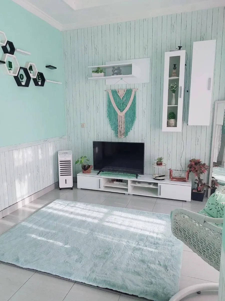 Interior ruang TV minimalis dengan kabinet putih, rak dinding heksagonal, dan karpet bulu biru muda yang lembut.