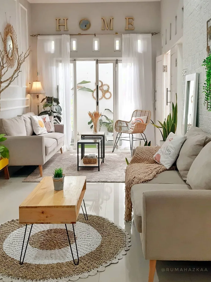 Desain ruang tamu terbuka dengan dekorasi dinding bertuliskan HOME, kursi rotan, dan akses langsung ke taman belakang yang asri.