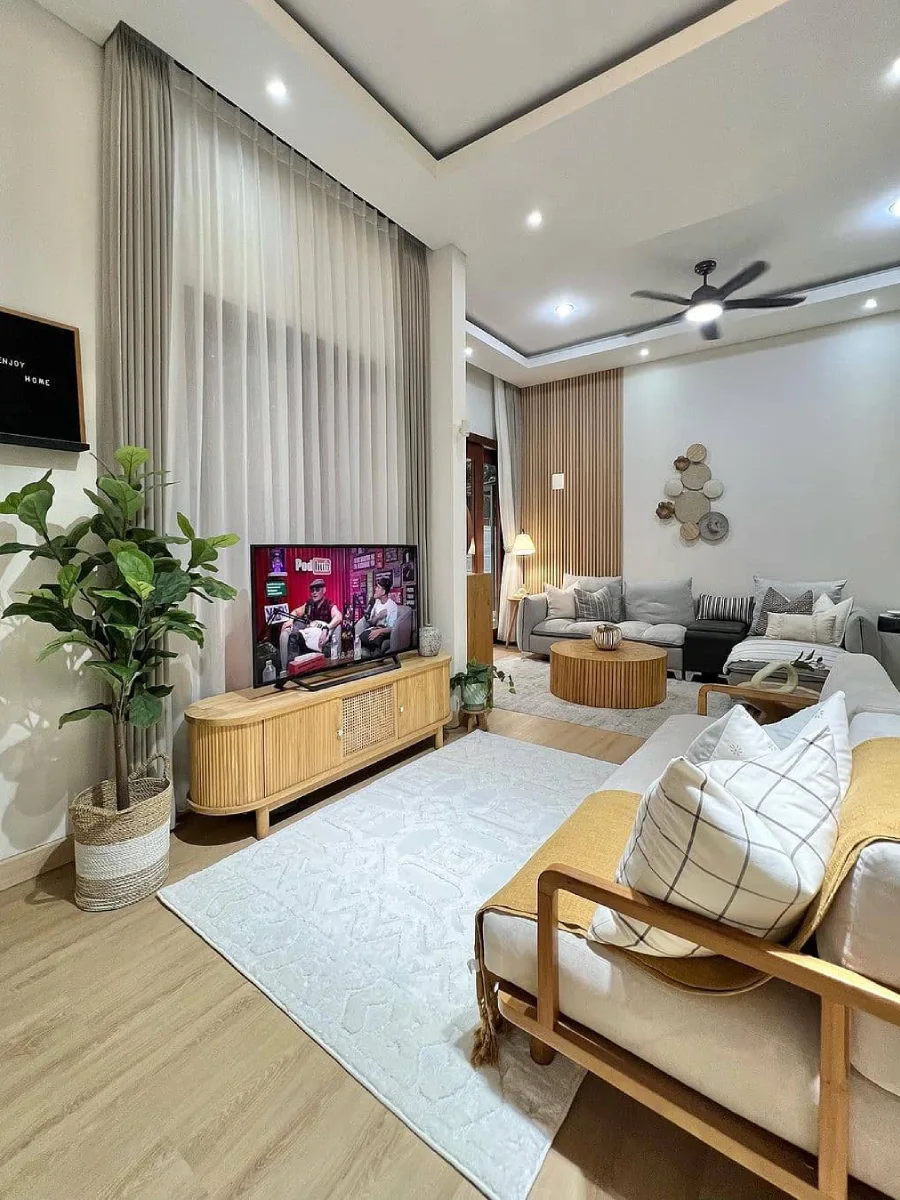 Ruang keluarga nyaman dengan sofa L abu-abu, dinding panel kayu minimalis, dan jendela besar dengan gorden putih transparan.