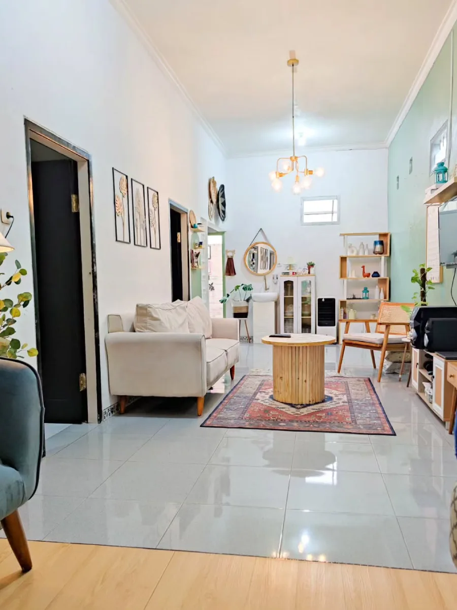 Ruang keluarga mungil memanjang dengan dinding hijau pastel, meja kayu bundar, dan sentuhan dekorasi bergaya bohemian.