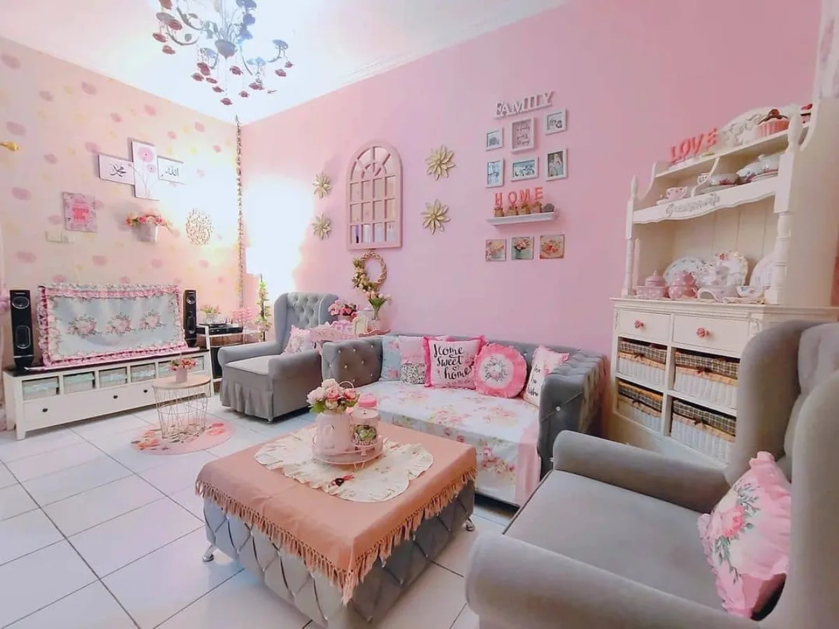 Sudut ruang keluarga bertema feminin dan romantis dengan dominasi warna pink, menampilkan lemari putih bergaya vintage dengan keranjang rotan, kursi lengan (armchair) abu-abu yang nyaman, serta berbagai hiasan dinding yang tertata rapi.