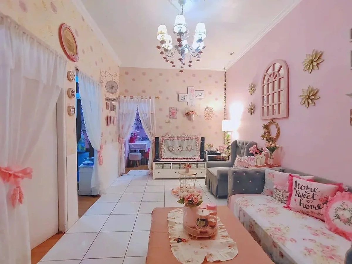 Ruang keluarga dengan desain interior shabby chic bernuansa pink, menampilkan sofa dengan bantal 'Home Sweet Home', dinding merah muda dengan dekorasi bunga emas, lampu gantung kristal yang elegan, dan lantai keramik putih yang memberikan kesan bersih.
