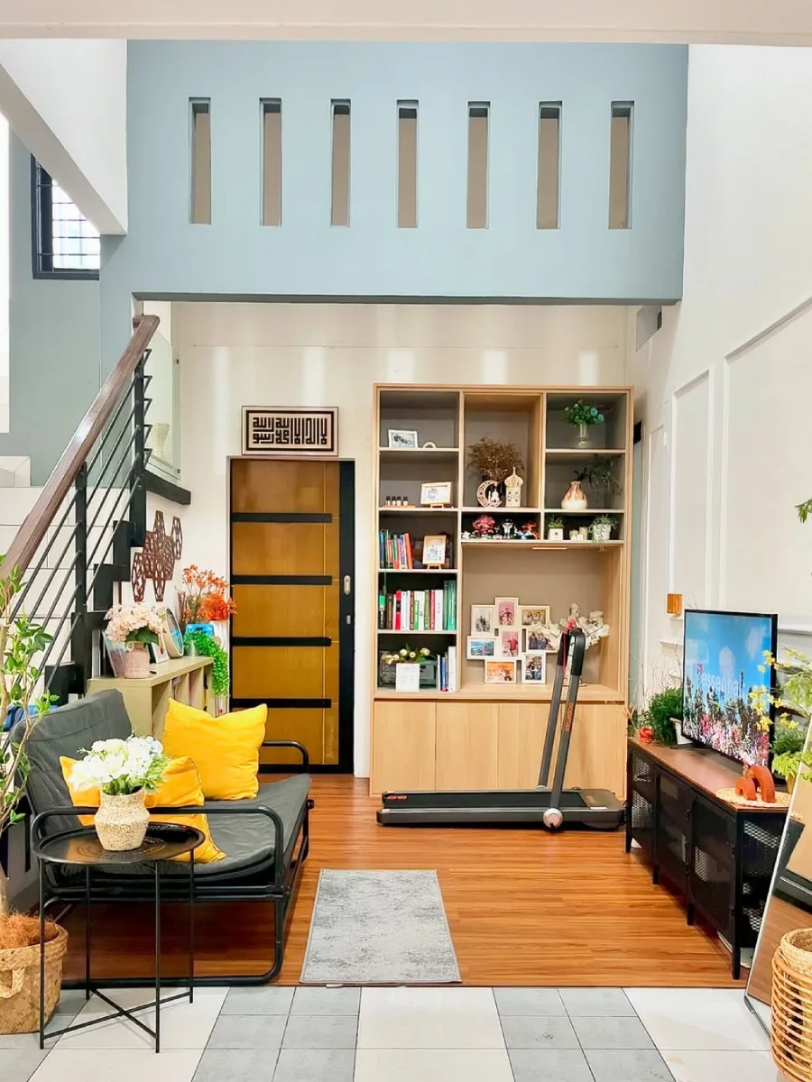 Interior ruang keluarga dengan lantai kayu, rak buku besar, treadmill portable, dan sofa abu-abu dengan bantal kuning yang cerah.